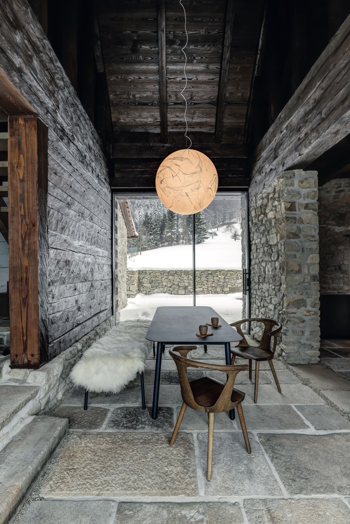  

Lustr Moon od italského designéra Davida Groppiho sehrál v celém projektu zásadní roli. Od začátku bylo jasné, že je pro tento prostor jako dělaný. Jídelní stůl, lavice a stolek v kuchyni jsou od přední české firmy Todus. Každý stůl má jinou tloušťku materiálu podle účelu: jídelní čtyři a pracovní v sousední kuchyni dvanáct milimetrů. Oba jsou vyrobené novou technologií tažení keramiky, která se používá především na obklady a dlažbu, protože umožňuje realizaci velkých ploch beze spár. Křesílka jsou z kolekce In Between od &Tradition. Prosklení domu na obě strany vytváří nepřerušený tok pohybu a světla. Interiér zevnitř ven propojují také kamenné bloky na podlaze: v létě se počítá s jejich využitím hned na třech místech – na výhled do údolí je připravena ranní snídaňová terasa, u kamenného štítu nasměrovaného k lesu bude posezení ideální na oběd a na jihozápadě zase do výseku lesa zapadá slunce. V původním průjezdu a kuchyni jsou okna z tenkých hliníkových profilů a v obytné části z modřínu, aby bylo původní rozdělení stále znatelné. Rozmístění oken i dveří je originální, dalo se snadno odečíst ze záseků ve dřevě.
