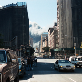 Vpředu zdánlivě klidná ulice, na pozadí lze vidět valící se oblaka popela a prachu ze Světového obchodního centra.