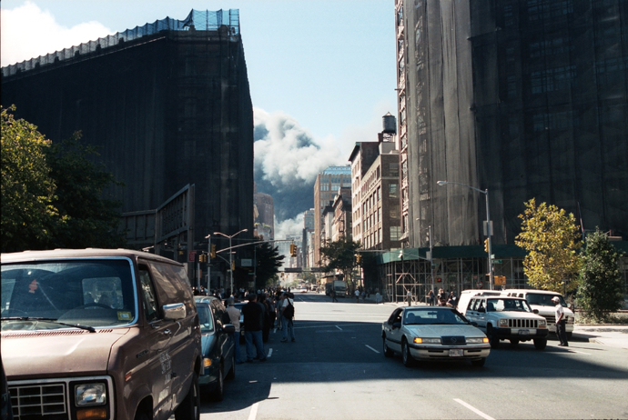 Vpředu zdánlivě klidná ulice, na pozadí lze vidět valící se oblaka popela a prachu ze Světového obchodního centra.