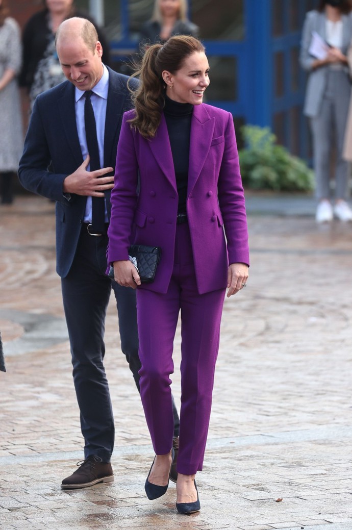 Kate Middleton ve fialovém kalhotovém kostýmku, který zkombinovala s černými lodičkami, černým prošívaným psaníčkem a kruhovými náušnicemi.

Pokud byste si na českých e-shopech chtěla pořídit podobný outfit, jeho celková cena by činila zhruba 16 241 Kč.