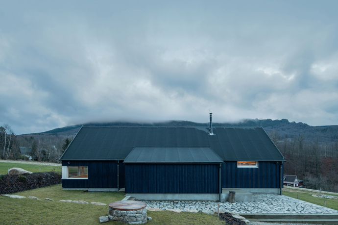 Modrý šindelový obklad a elegantní bílé rámy oken vytvářejí kontrast, který nemůžete přehlédnout. Architektka Markéta Cajthamlová a její tým se postarali o každý detail, ať už jde o interiér plný dřeva nebo promyšlenou dispozici, která nabídne vše, co potřebujete.
