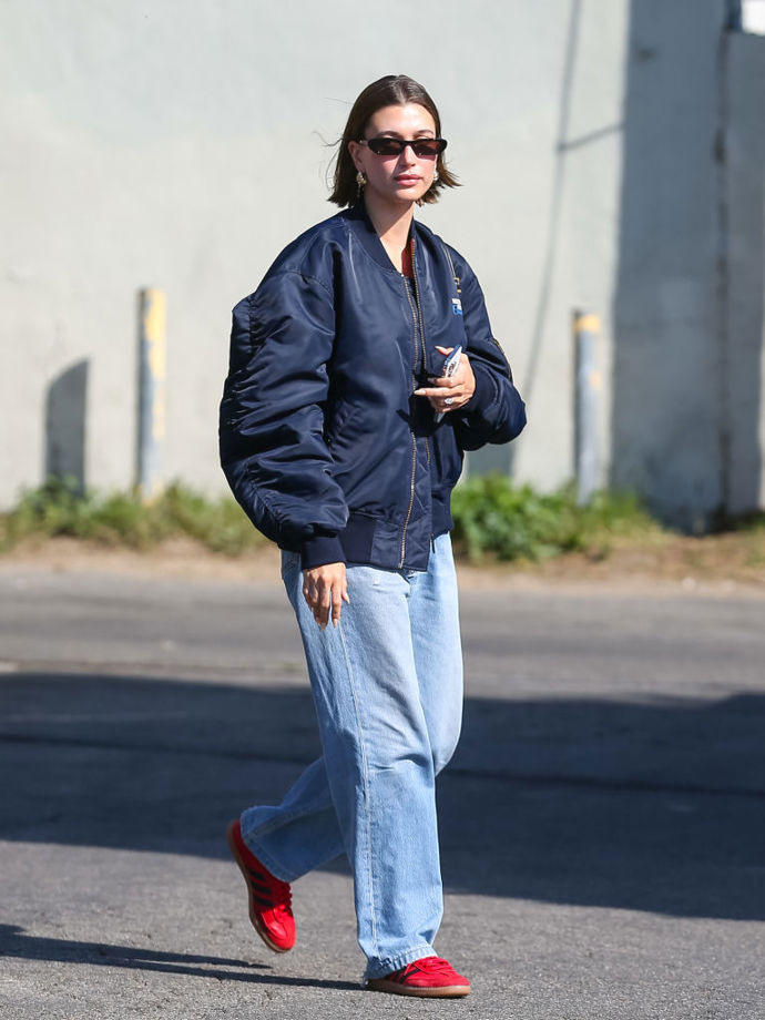 Hailey Bieber v ležérním outfitu v podobě oversized bomberu a džínů doplněném červenými trendy teniskami.
