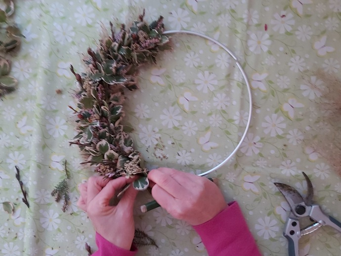 Drátkem větvičku připevníme cca 1 cm od konce jejího stonku tak, že drátek obtočíme kolem rostlinky od spodu přes její stonek horem do vnitřku kruhu a protáhneme zpět. 

