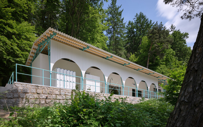Návrh vypracovalo architektonické studio Mjölk architekti a zahrnovalo například kombinaci CLT konstrukce s lehkým dřevěným rámem, lehké trámkové konstrukce, a to vše pod tyrkysovou střechou, která slouží jako ochrana i stínidlo. Když na ni padne odpolední světlo, odráží se od ní paprsky jako od vodní hladiny. 
