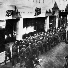 Němečtí vojáci pochodují před lékárnou na Normanských ostrovech, 40. léta 20. století. Claude Cahun se spolu se svou partnerkou Marcel Moore účastnila protinacistického odboje.
