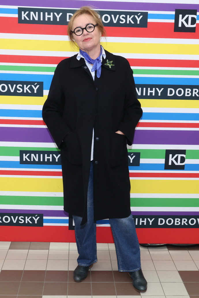 Jitka Sedláčková kombinuje černý kabát s modrým šátkem a brýlemi s kulatými obroučkami. 