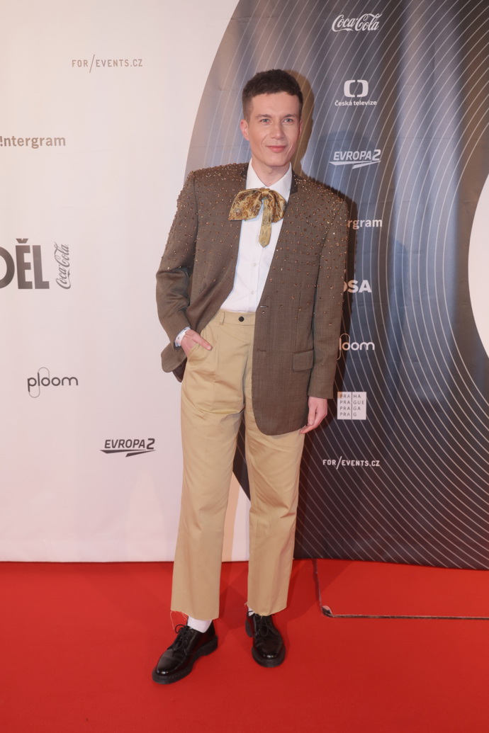 Herec Daniel Krejčík v bílé košili, béžových kalhotách a hnědém sáčku. Nechybí efektní vázanka pod krkem. 