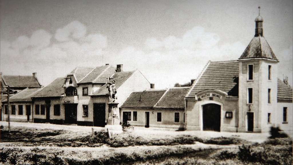 Původní hasičárna stojící na hlavní ulici byla menší, ale u vjezdu měla architektonicky elegantní oblouk. Socha před ní stojí dodnes.
