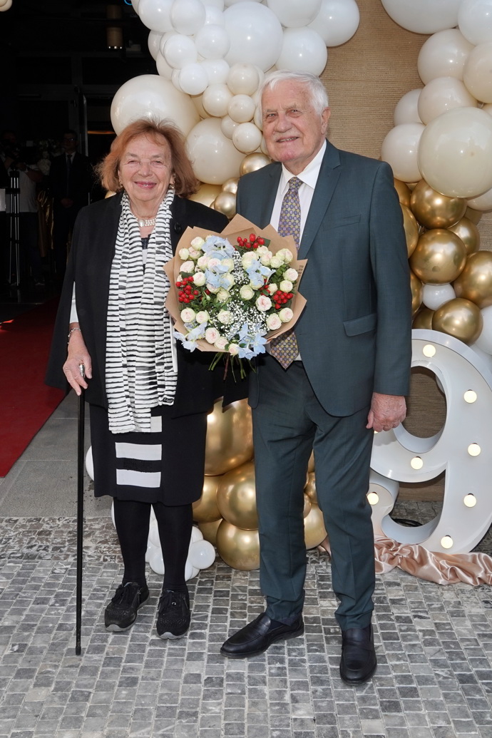 Jubileum Jiřiny Bohdalové přišli oslavit i manželé Klausovi. Exprezident vsadila na šedozelený oblek s bílou košilí a ladící vzorovanou kravatou  s nádechem béžové, bývalá první dáma elegantní černobílou klasiku propojila s pohodlnou obuví.