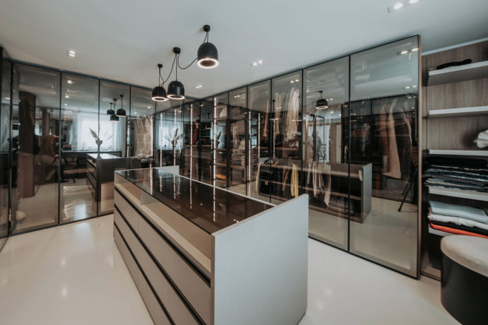 Prostorná šatna využívá kouřové sklo, tmavé rámy a centrální ostrůvek se zásuvkami, takže působí spíš jako butik než běžná vestavěná skříň