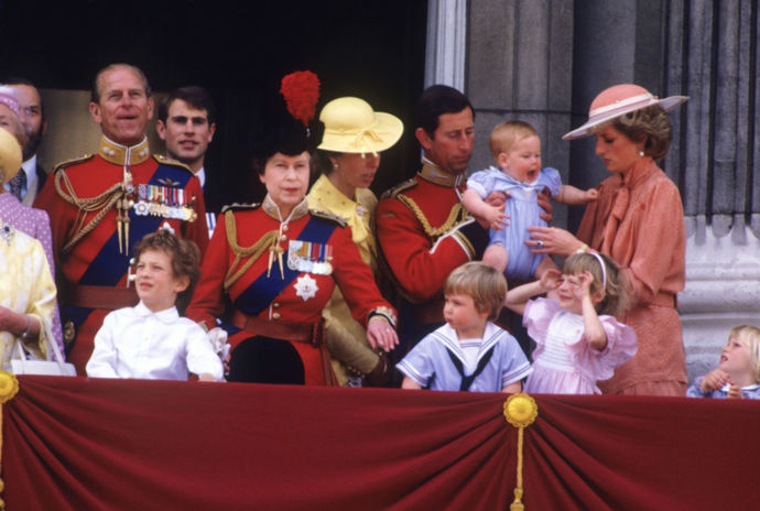 (zleva) Princ Philip, vévoda z Edinburghu, princ Edward, královna Alžběta II., princezna Anna, princ Charles držící prince Harryho a princezna Diana v broskvových šatech s motýlkem od Jana Van Veldena a v klobouku od Fredericka Foxe v roce 1985, v první řadě malý princ William