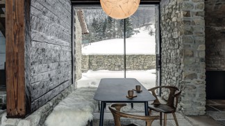 

Lustr Moon od italského designéra Davida Groppiho sehrál v celém projektu zásadní roli. Od začátku bylo jasné, že je pro tento prostor jako dělaný. Jídelní stůl, lavice a stolek v kuchyni jsou od přední české firmy Todus. Každý stůl má jinou tloušťku materiálu podle účelu: jídelní čtyři a pracovní v sousední kuchyni dvanáct milimetrů. Oba jsou vyrobené novou technologií tažení keramiky, která se používá především na obklady a dlažbu, protože umožňuje realizaci velkých ploch beze spár. Křesílka jsou z kolekce In Between od &Tradition. Prosklení domu na obě strany vytváří nepřerušený tok pohybu a světla. Interiér zevnitř ven propojují také kamenné bloky na podlaze: v létě se počítá s jejich využitím hned na třech místech – na výhled do údolí je připravena ranní snídaňová terasa, u kamenného štítu nasměrovaného k lesu bude posezení ideální na oběd a na jihozápadě zase do výseku lesa zapadá slunce. V původním průjezdu a kuchyni jsou okna z tenkých hliníkových profilů a v obytné části z modřínu, aby bylo původní rozdělení stále znatelné. Rozmístění oken i dveří je originální, dalo se snadno odečíst ze záseků ve dřevě.
