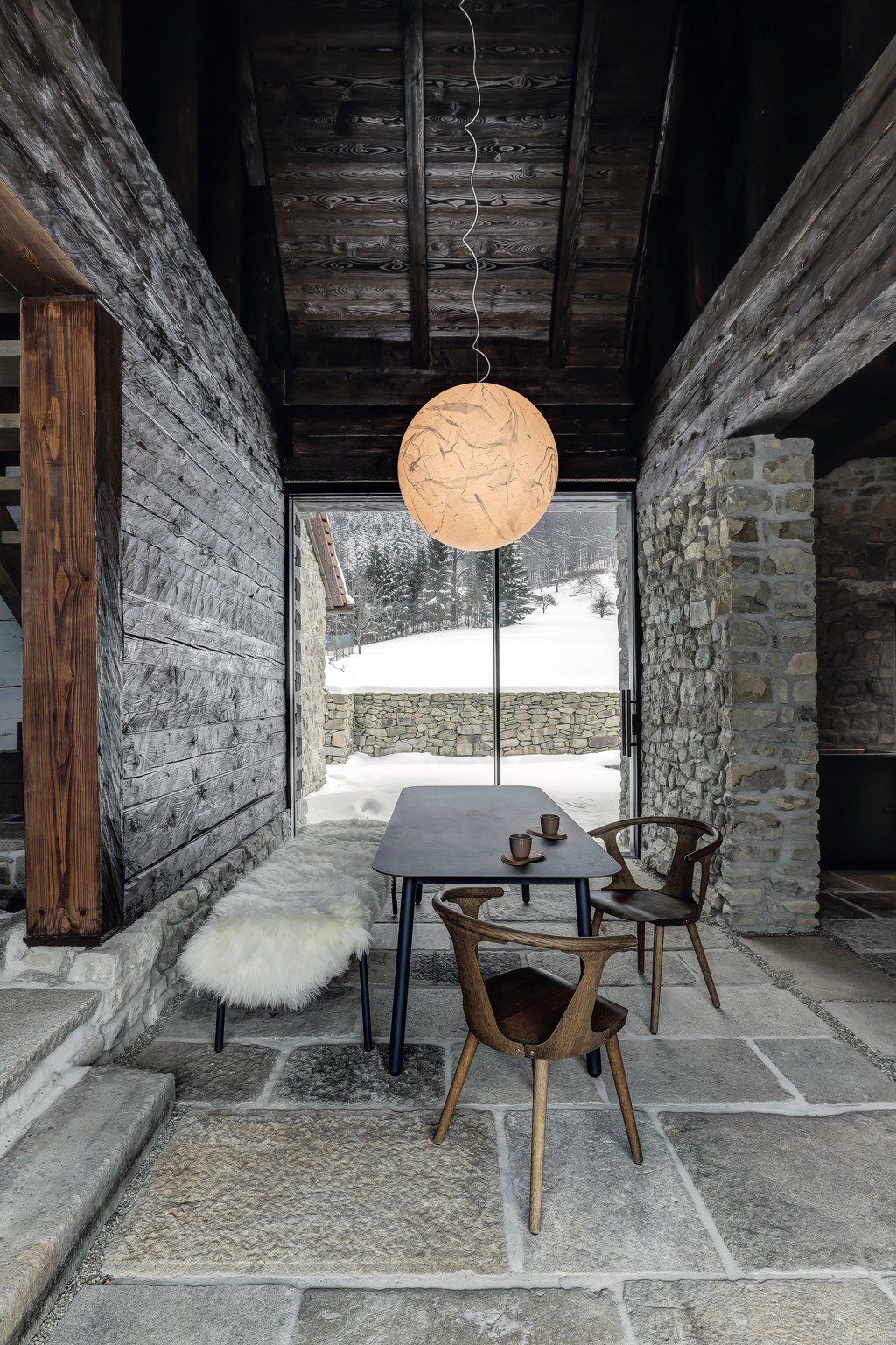 Lustr Moon od italského designéra Davida Groppiho sehrál v celém projektu zásadní roli. Od začátku bylo jasné, že je pro tento prostor jako dělaný. Jídelní stůl, lavice a stolek v kuchyni jsou od přední české firmy Todus. Každý stůl má jinou tloušťku materiálu podle účelu: jídelní čtyři a pracovní v sousední kuchyni dvanáct milimetrů. Oba jsou vyrobené novou technologií tažení keramiky, která se používá především na obklady a dlažbu, protože umožňuje realizaci velkých ploch beze spár. Křesílka jsou z kolekce In Between od &Tradition. Prosklení domu na obě strany vytváří nepřerušený tok pohybu a světla. Interiér zevnitř ven propojují také kamenné bloky na podlaze: v létě se počítá s jejich využitím hned na třech místech – na výhled do údolí je připravena ranní snídaňová terasa, u kamenného štítu nasměrovaného k lesu bude posezení ideální na oběd a na jihozápadě zase do výseku lesa zapadá slunce. V původním průjezdu a kuchyni jsou okna z tenkých hliníkových profilů a v obytné části z modřínu, aby bylo původní rozdělení stále znatelné. Rozmístění oken i dveří je originální, dalo se snadno odečíst ze záseků ve dřevě.
Lustr Moon od italského designéra Davida Groppiho sehrál v celém projektu zásadní roli. Od začátku bylo jasné, že je pro tento prostor jako dělaný. Jídelní stůl, lavice a stolek v kuchyni jsou od přední české firmy Todus. Každý stůl má jinou tloušťku materiálu podle účelu: jídelní čtyři a pracovní v sousední kuchyni dvanáct milimetrů. Oba jsou vyrobené novou technologií tažení keramiky, která se používá především na obklady a dlažbu, protože umožňuje realizaci velkých ploch beze spár. Křesílka jsou z kolekce In Between od &Tradition. Prosklení domu na obě strany vytváří nepřerušený tok pohybu a světla. Interiér zevnitř ven propojují také kamenné bloky na podlaze: v létě se počítá s jejich využitím hned na třech místech – na výhled do údolí je připravena ranní snídaňová terasa, u kamenného štítu nasměrovaného k lesu bude posezení ideální na oběd a na jihozápadě zase do výseku lesa zapadá slunce. V původním průjezdu a kuchyni jsou okna z tenkých hliníkových profilů a v obytné části z modřínu, aby bylo původní rozdělení stále znatelné. Rozmístění oken i dveří je originální, dalo se snadno odečíst ze záseků ve dřevě.