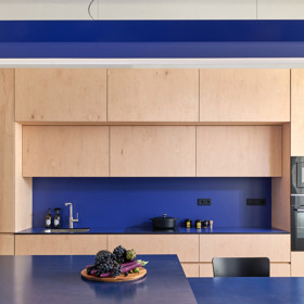 Světlou kuchyňskou sestavu z překližky doplňuje sytě modrá pracovní plocha a ostrůvek, který plynule navazuje na jídelní stůl