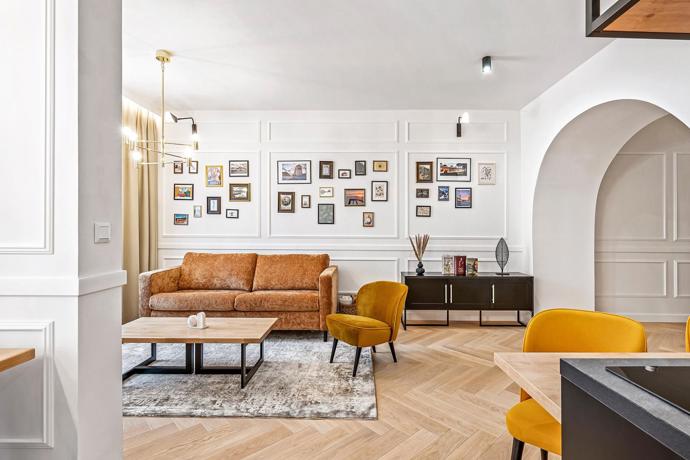 Galerijní stěna, koženě působící sedačka a hořčicová křesla dodávají jinak světlému interiéru osobnější a méně hotelový charakter