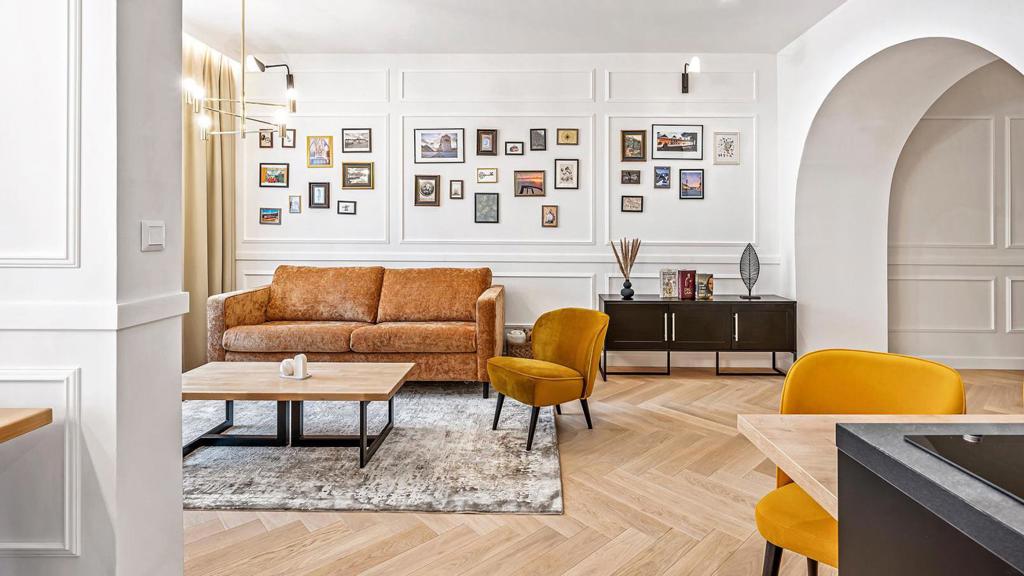 Galerijní stěna, koženě působící sedačka a hořčicová křesla dodávají jinak světlému interiéru osobnější a méně hotelový charakter Galerijní stěna, koženě působící sedačka a hořčicová křesla dodávají jinak světlému interiéru osobnější a méně hotelový charakter
