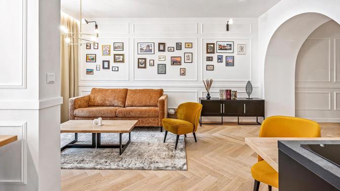 Galerijní stěna, koženě působící sedačka a hořčicová křesla dodávají jinak světlému interiéru osobnější a méně hotelový charakter